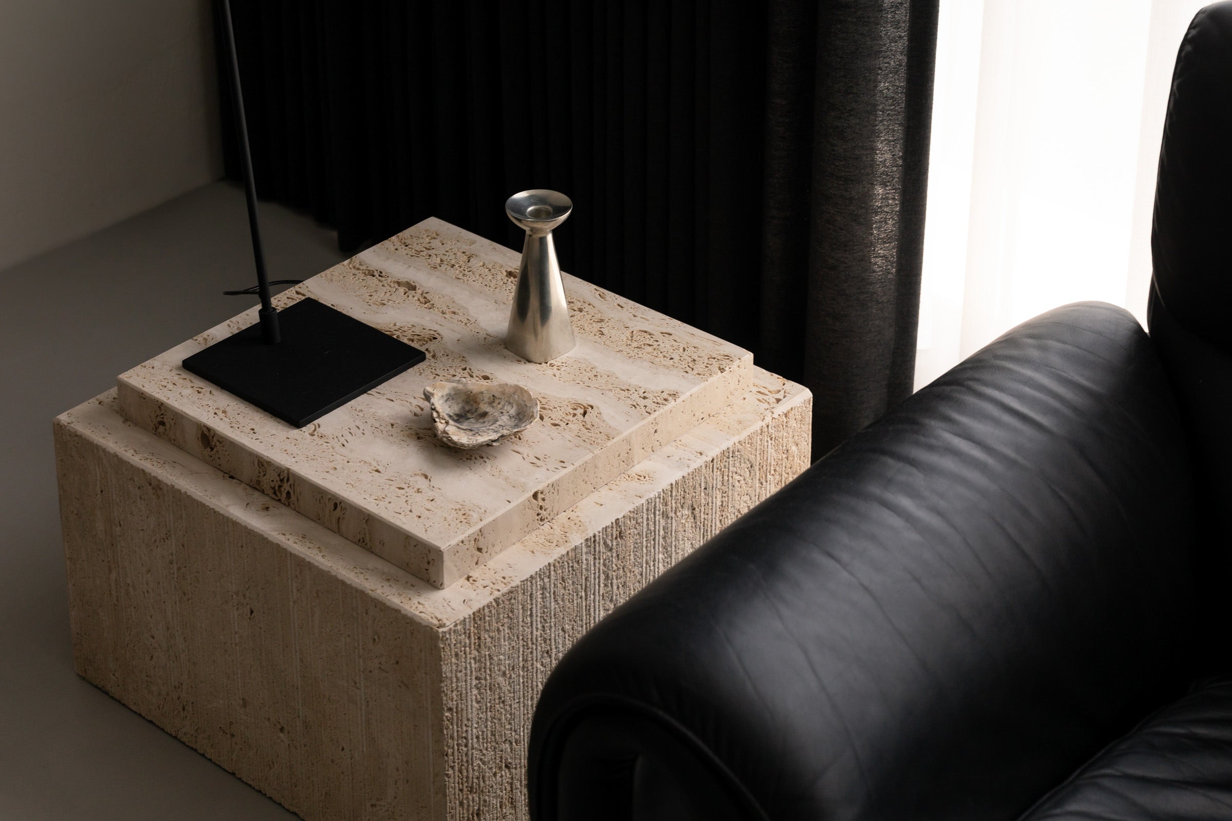 Un'common Bari travertine coffee table beside a black leather sofa, set against a dark accent wall and sheer curtain.