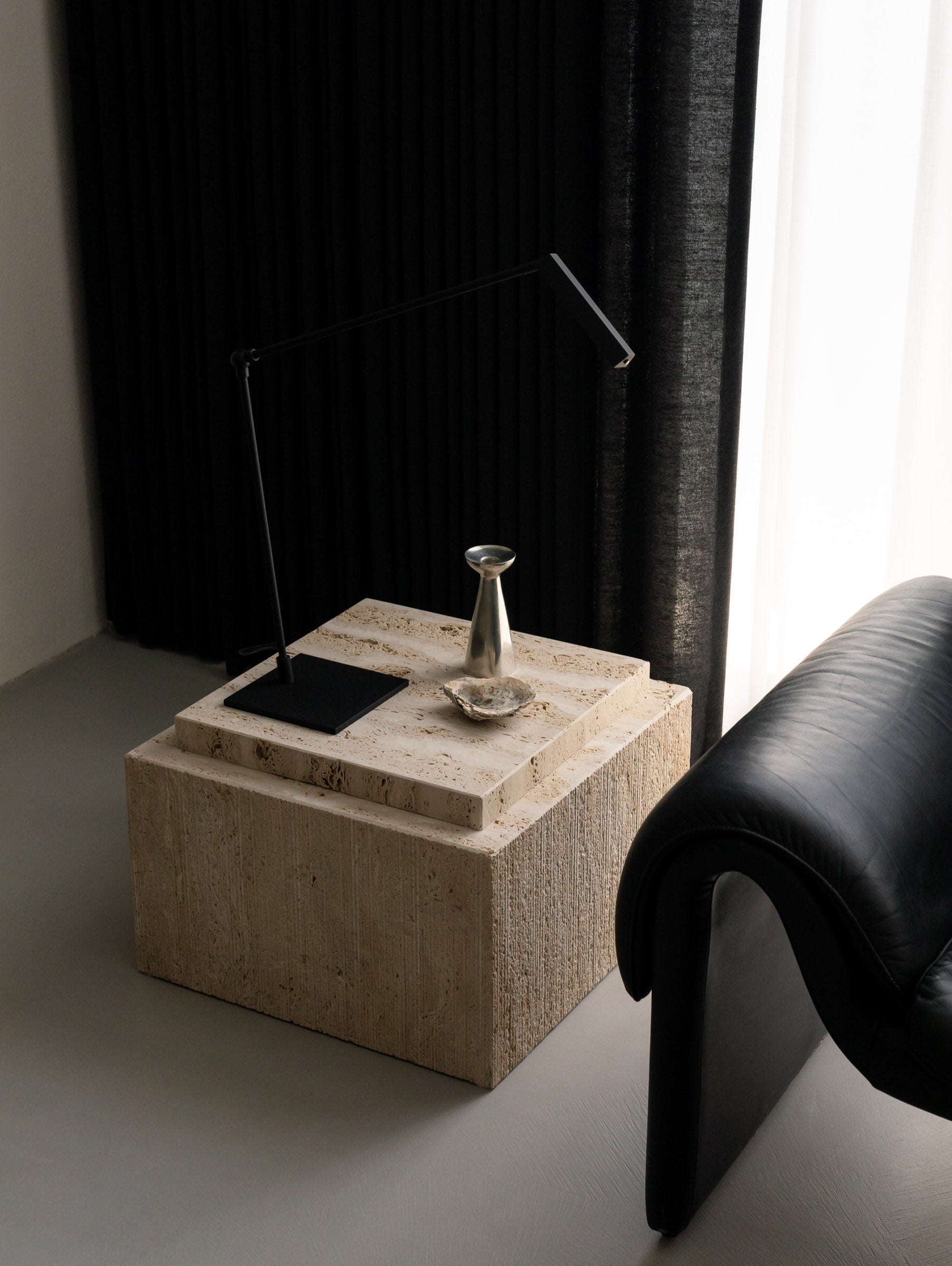 Close-up of an Un'common BARI travertine coffee table styled with a metal candle holder and a small object, beside a black leather chair and a dark curtain