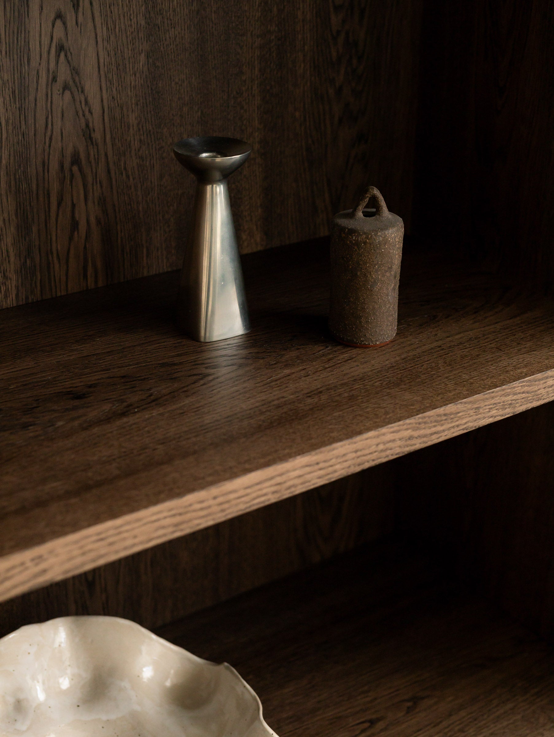 Close-up of a brushed metal candle holder and a small textured ceramic vessel on a shelf of the Un'common BLOCK cabinet, with a white organic bowl visible below.