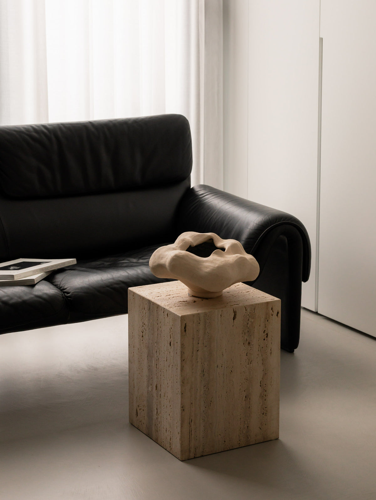 Un'common COI travertine pedestal styled with an organic cream ceramic vessel, beside a black leather sofa with stacked books."