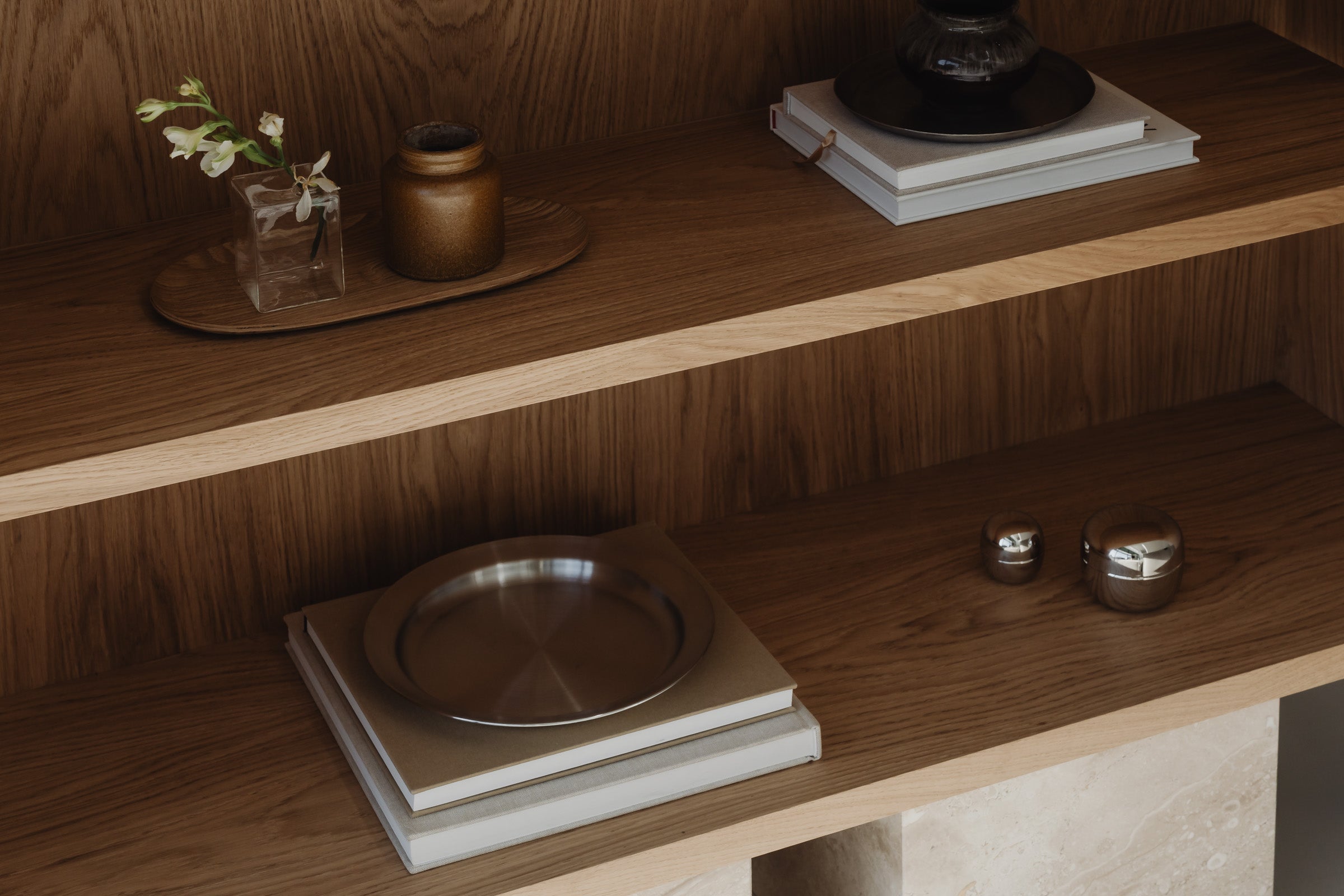 Un'common Sideboard unit in warm oak, styled with a glass bud vase, a stoneware jar, a wooden tray, stacked books, a ceramic lamp base, a steel plate, and small metallic trinket boxes.