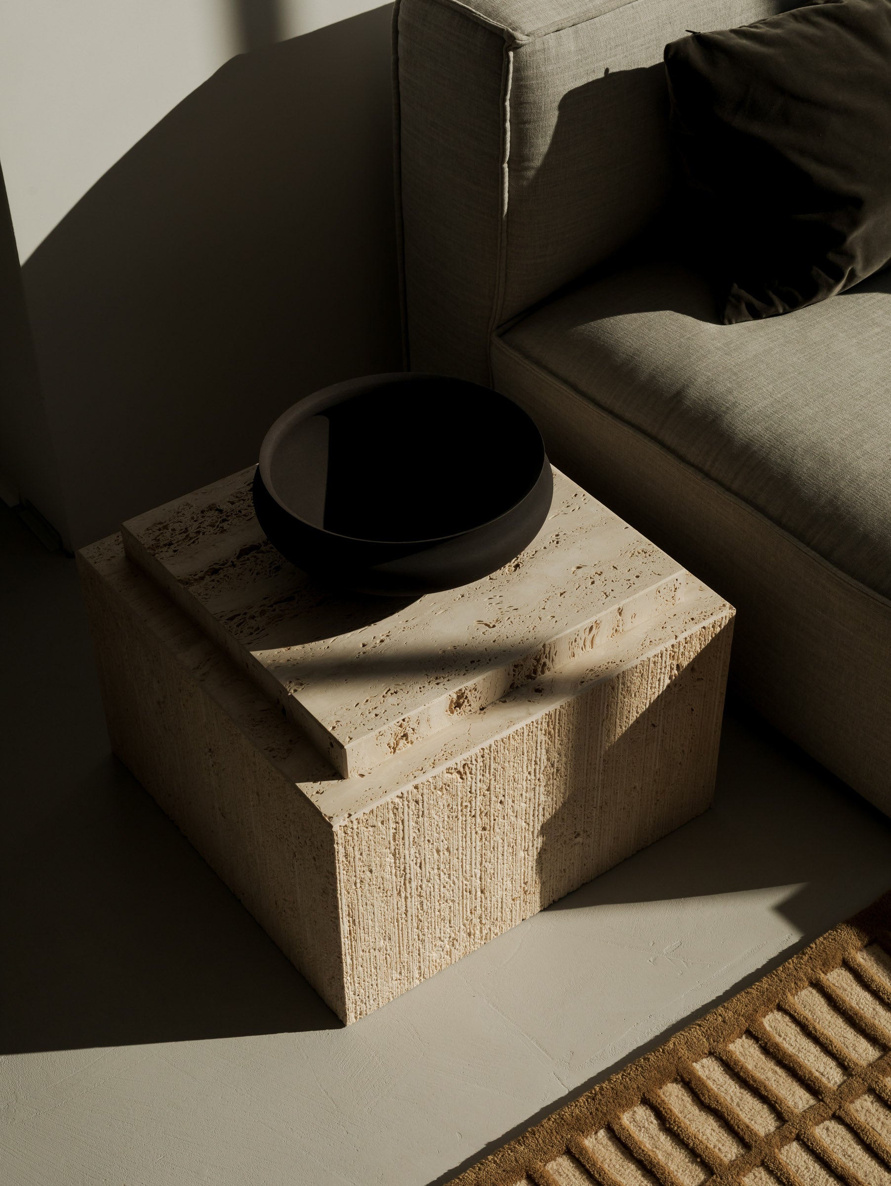 Close-up of an Un'common BARI travertine coffee table with a black ceramic bowl, beside a beige sofa and a woven cane rug."

